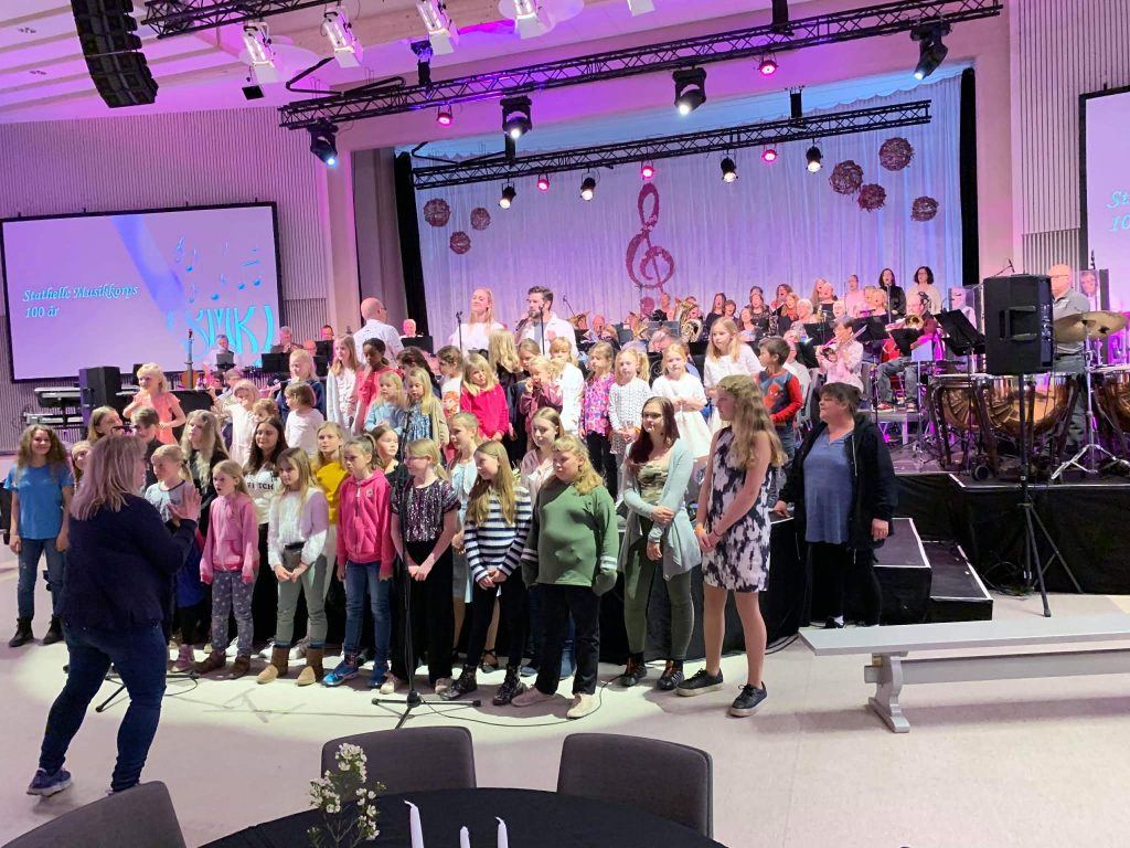 barnekor og blanda voksen kor med solister på en scene, med Ingrid som dirigent