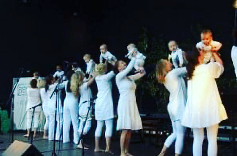 Flere kvinner som løfter babyen sin i været, alle er kledd i hvit og står på en scene 