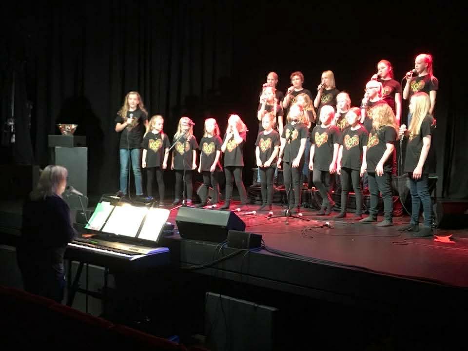 barnekor på en scene, de er kledd i like klær av sort farge, det er rødt lys på scenen.