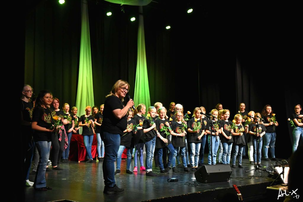 Ingrid som snakker i mikrofon på en scene, bak henne står barnekor i like klær